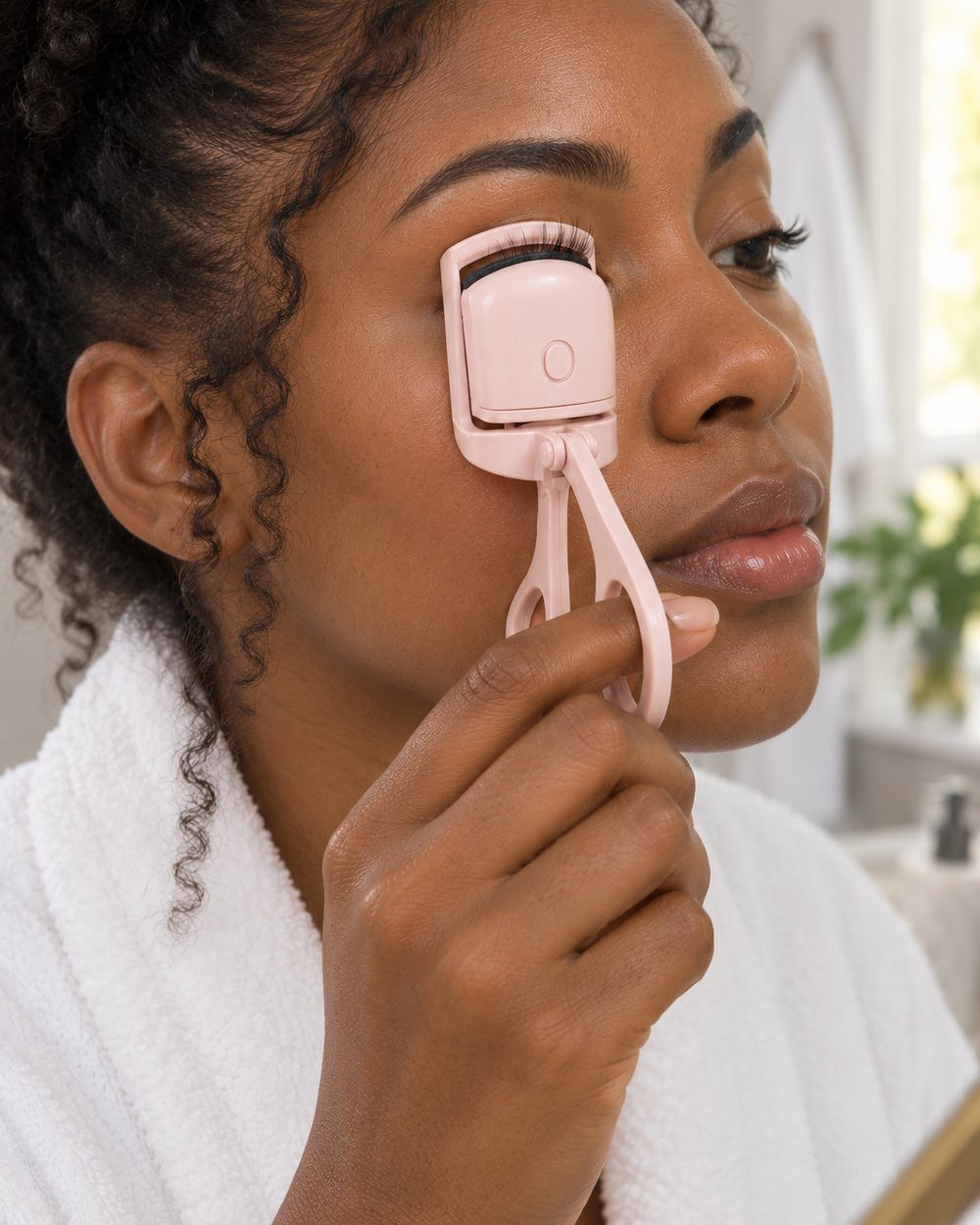Woman clamping the heated curler on her upper lashes in front of a bathroom mirror