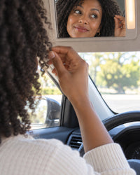 A woman checks frizzy flyaways around her face in a car mirror
