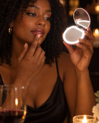 Woman checking her lipstick in the lit compact mirror at a dim restaurant table