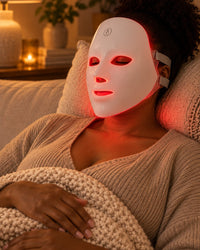 Woman lying back on a sofa wearing the white LED mask, glowing soft red, eyes closed