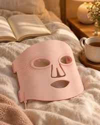 Pink silicone LED mask on a linen-covered bed beside a cup of tea and a book